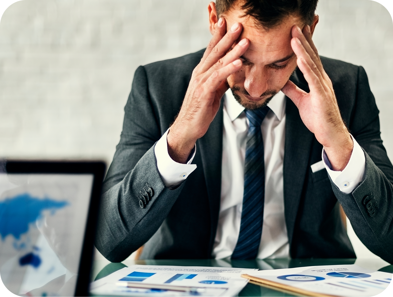A man in a suit sitting at a desk with papers and graphs, holding his head in his hands, appearing stressed or contemplative.